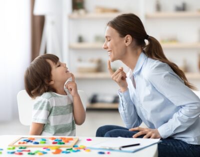Speech training for kids. Professional woman specialist training with little boy at cabinet, teaching him right articulation exercises, side view